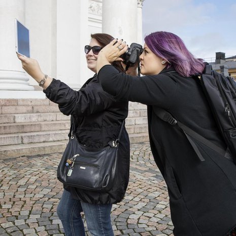 Näin syntyy blogipostaus.  Satu pitelee tablettia, jolla näkyy pysäytyskuva elokuvasta.  Tiia ottaa kuvan, jossa tabletin kuva istuu täydellisesti taustaan.